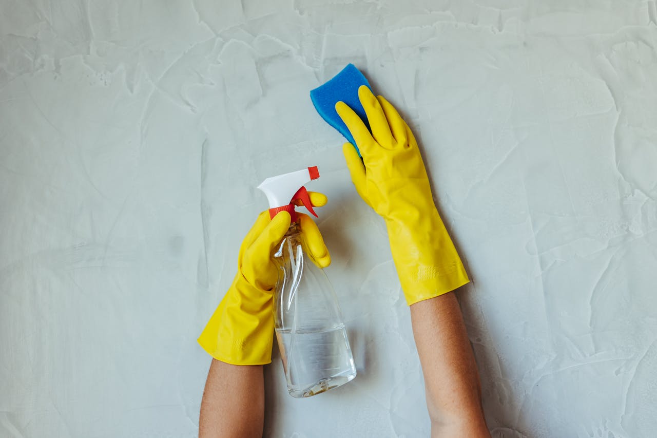 hero-img Close-up of hands in yellow gloves cleaning a wall with a spray bottle and sponge.
